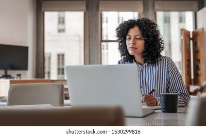 Young female entrepreneur using a laptop and taking notes while working from home at her dining table - Shutterstock ID 1530299816