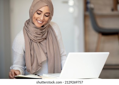 Young female entrepreneur wearing a hijab sitting at a desk in her home office working online with a laptop. Muslim woman working in home office. She seems to be concentrated and glad