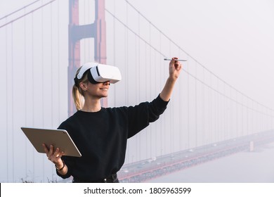 Young female extend hand with digital pencil for interactive touch over air while standing in against bridge construction wearing virtual reality headset. Hipster girl designing pen copy space with VR - Powered by Shutterstock