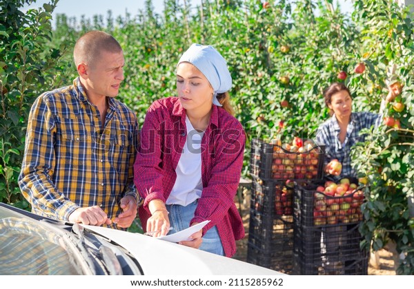 Young female\
farmer discussing contract for transportation of apple harvest with\
representative of transport company outdoors against background of\
farm orchard in\
summertime