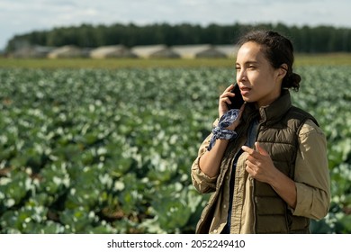 Young female farmer or owner of farm speaking on mobile phone against plantation