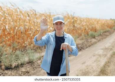 A young female farmer is smiling and waving while standing in a sunlit cornfield. She wears a casual denim shirt and a cap, embodying the simplicity and beauty of rural life - Powered by Shutterstock