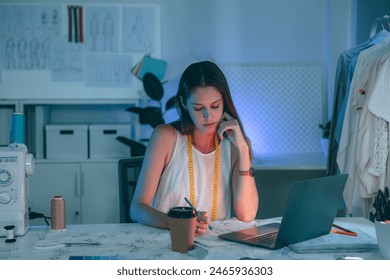 Young female fashion designer working late at night in her studio, focused on laptop and pattern sketches. Creative workspace environment. - Powered by Shutterstock