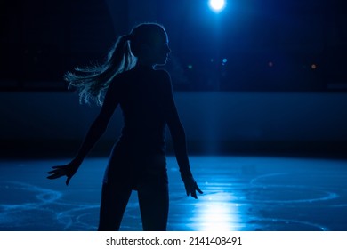 Young Female Figure Skater Is Performing Woman's Single Skating Choreography On Ice Rink. Silhouette Of Woman Practicing Skills On Ice Arena Against Background Of Blue Light And Spotlights. Close Up.