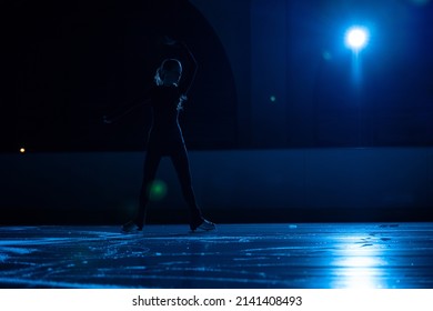 Young Female Figure Skater Is Performing Woman's Single Skating Choreography On Ice Rink. Dark Silhouette Of Woman Practicing Skills On Ice Arena Against Background Of Soft Blue Light And Spotlights.