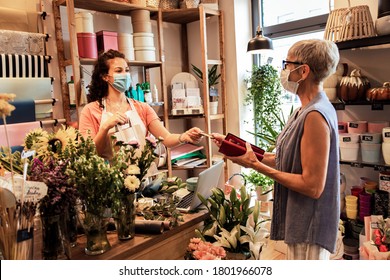 Young Female Florist With Mask Working In Flower Shop, Selling Flower Arrangement To Senior Woman.