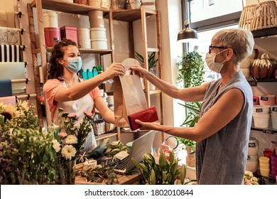 Young Female Florist With Mask Working In Flower Shop, Selling Flower Arrangement To Senior Woman.