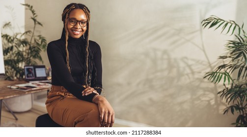 Young female freelancer smiling at the camera while working on a new project in her home office. Happy young photographer sitting on a chair with her creative desk in the background. - Shutterstock ID 2117288384