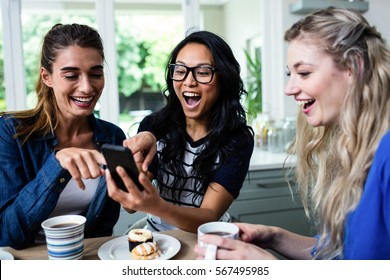 Young Female Friends Laughing While Pointing On Mobile Phone During Breakfast At Home