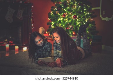 Young Female Friends Lying On Rug Beside Christmas Tree And Looking At The Tablet. Beautiful New Year Room Interior