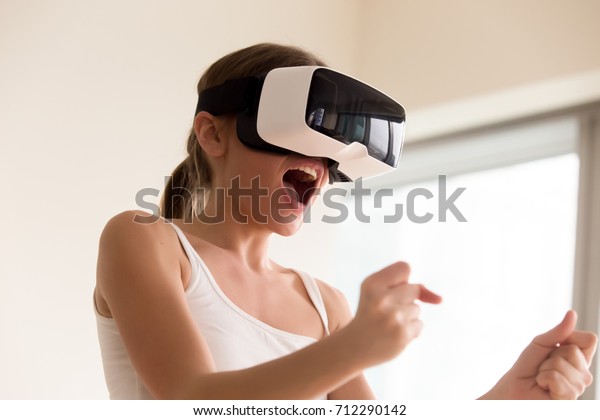 Young female gamer in wearable VR headset playing\
action 3d video game at home, excited teen holding wheel and\
screaming driving digital racing car in simulator application,\
virtual gaming concept