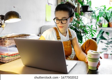 Young female gardener in glasses using laptop, communicates on internet with customer in home garden/greenhouse, hold reusable coffee/tea mug.Cozy office workplace, remote work, stay home concept