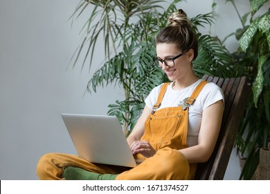 Young female gardener in glasses wearing overalls, sitting on wooden chair in greenhouse, using laptop after work, communicates on Internet with customer. Plant on background.Home gardening, freelance - Shutterstock ID 1671374527