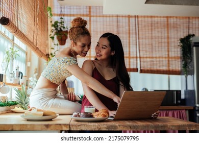 A Young Female Gay Couple Is At Home Using Their Computer. One Is Standing In Front Of The Computer, And The Other One I Sitting On The Kitchen Counter Top Pointing At It.