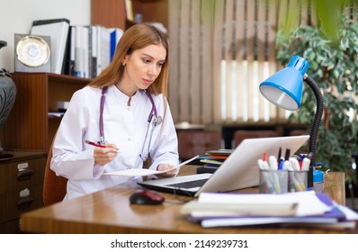 Young Female General Practitioner Working In The Clinic Studies The Patient's Outpatient Card, Writing Down The Appointment ..of Treatment, Sitting At The Workplace In The Office