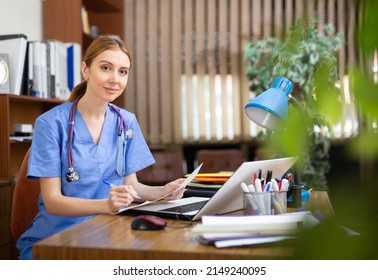 Young Female General Practitioner Working In The Clinic Studies The Patient's Outpatient Card, Writing Down The Appointment ..of Treatment, Sitting At The Workplace In The Office