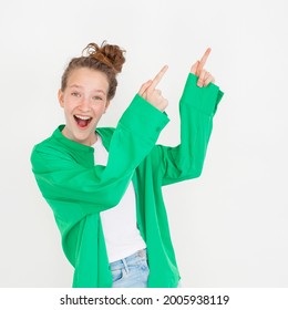 Young Female Girl With Happy Smile, Points Away With Both Hands, Standing Against White Background In Green Shirt. Portrait Of Fun Teen Student Studio Shot