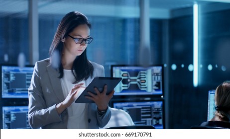 Young Female Government Employee Wearing Glasses Uses Tablet in System Control Center. In the Background Her Coworkers are at Their Workspaces with many Displays Showing Valuable Data.