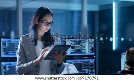 Young Female Government Employee Wearing Glasses Uses Tablet in System Control Center. In the Background Her Coworkers are at Their Workspaces with many Displays Showing Valuable Data.