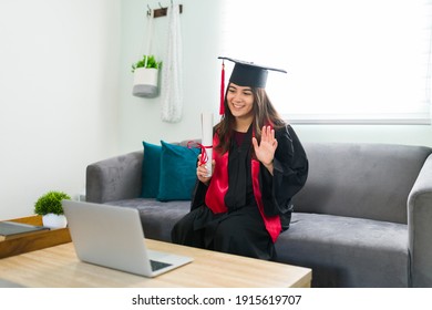 Young Female Graduate In A Black Gown Sitting On The Couch At Home And Waving During A Virtual Graduation Ceremony. Pretty Woman In Her 20s With A Face Mask Receiving Her College Diploma