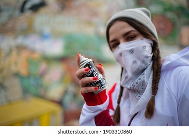 Young Female Graffiti Artist With Red Nails Stands Against Graffiti Wall With Her Face Covered In White Hoodie And Holds A Spray Can