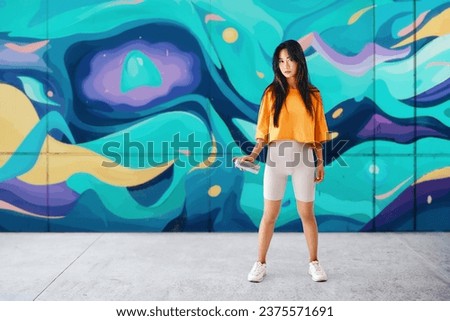 Young female graffiti painter standing near the wall with her paintings with copy space. Street art concept Full length portrait