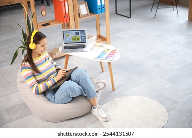 Young female graphic designer in headphones working with tablet on beanbag at office - Powered by Shutterstock