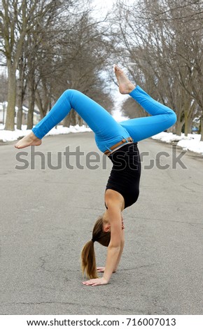 young female gymnast doing handstand in city street. snow cover and trees. she is wearing bright royal blue pants and black top. long pony tail touching the ground. 