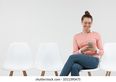 Young Female With Hair Bun Holding Generic Smart Phone, Checking Newsfeed On Social Media, Browsing Pictures  Looking At Screen With Happy And Cheerful Expression, Sitting In The Line