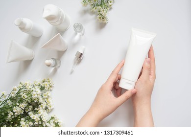 Young female hand holding blank white squeeze bottle plastic tube w/ organic natural skincare products and flower on white table. Packaging of lotion, cream or serum. Beauty cosmetic skincare concept.