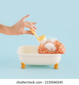 Young female hand washing brain with brush in tub with foam, on isolated pastel blue background. Minimal abstract concept of mental health, brain fog or brainwashing. Human head healthcare treatment.