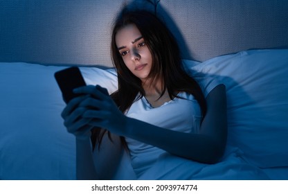 Young Female Having Insomnia Browsing Social Media Via Mobile Phone While Resting In Soft Bed In Dark Bedroom Late In Evening