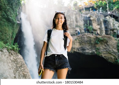 Young Female Hiker With Mobile Phone In Hand Looking At Camera And Enjoying Trip While Standing Among Rocky Cliffs Behind Waterfall