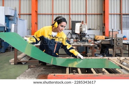 Young female hispanic labor dressed in polyester jacket uniform and ear muff working in metal sheet manufacture. Various kind of industrial machine are in the working area.