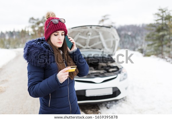 Young female holding road assistance membership\
or business card and call for help next to broken car with opened\
hood. Winter season.