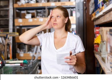 Young female holding shop list and choosing  tools at shelves in build store