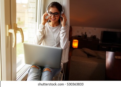 Young female at home.She sitting in her living room by the window and listening to music on laptop.