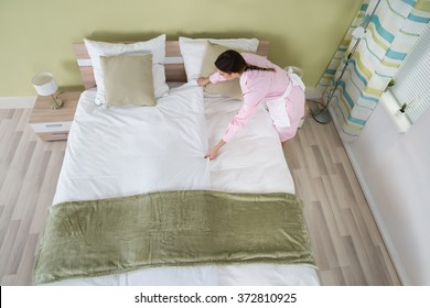 Young Female Housekeeper Arranging Bedsheet On Bed In Room