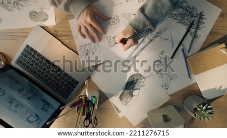 Young female illustrator drawing pencil sketches on paper. Drawings on the sheet of papers in front of her. Illustration animation, storyboard, video game film production, animation design studio.