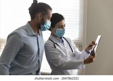 Young female Indian doctor in facemask showing xray screening images to male patient, explaining spinal injury, bone disease treatment, giving consultation after radiography scanning. Medic care
