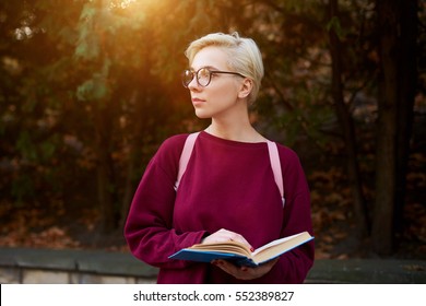 Young female international student spending time outdoor in park holding adventure novel, dreaming about her future trips,feeling pensive after reading on blurred background copy space for advertising