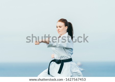 Young female karate fighter hitting a left punch. She is wearing black belt and white kimono. She is combing a pony tail. She is redhead and freckles. Blue sky and sea with bokeh effect on background