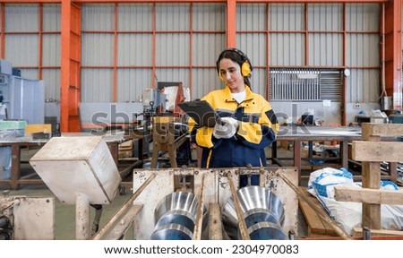 Young female labor dressed in polyester jacket uniform and ear muff reading document on clipboard, stand in front of steel and metal processing machinery. Atmosphere in metal sheet factory.