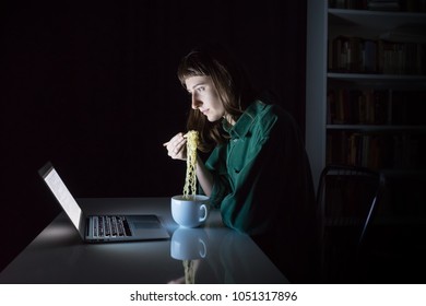 Young female at laptop computer eats instant ramen noodles late in the evening. Woman studying online overtime at night has fast dinner - concept of consuming unhealthy junk food at workplace