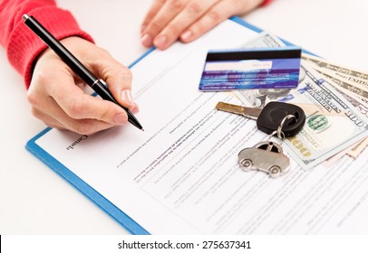 Young female leasing consultant signing car insurance contract in the office. Closeup of sold vehicle key with credit card and cash. Shallow depth of field.  