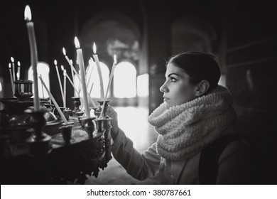 Young Female Lighting Candles In A Church During Praying.Yellow Votive Candles Burning.Woman Praying To God At St. Alexander Nevsky Cathedral.Christianity.Strong Christian Religion Faith