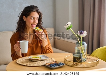 Young female looking through window while having breakfast by kitchen table served with sandwiches, blueberries and nuts on plates