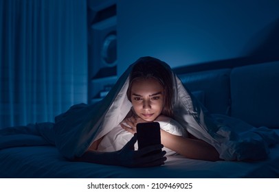 Young female lying under blanket on bed and watching horror movie on mobile phone in dark bedroom in late evening