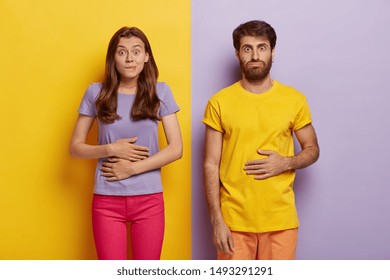 Young female and male touch stomaches, feel satiety after eating hearty meal, bites lips, have good appetite, dressed in casual outfit, pose against yellow and purple wall. Pleasant feeling in belly