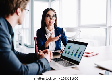 Young female manager talking to colleagues gesture and smiling having consultancy meeting about business, positive woman in formal wear asking question while man sitting near laptop with infographic